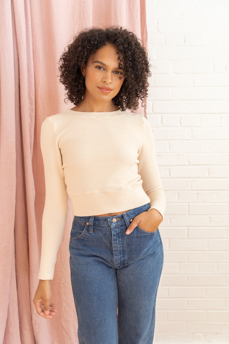 Woman wearing a beige sweater and blue jeans standing against a pink curtain and white brick wall.