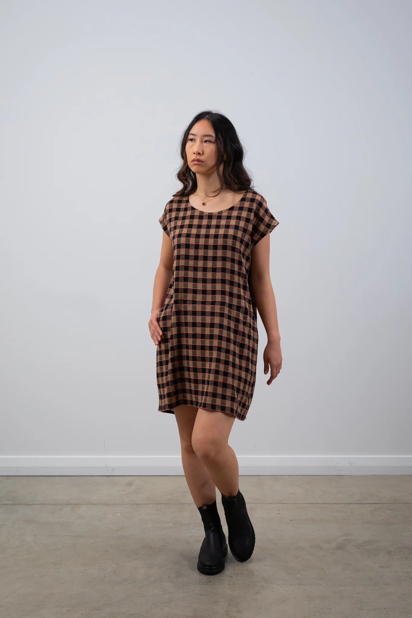 Woman wearing a brown checkered dress standing against a white wall.