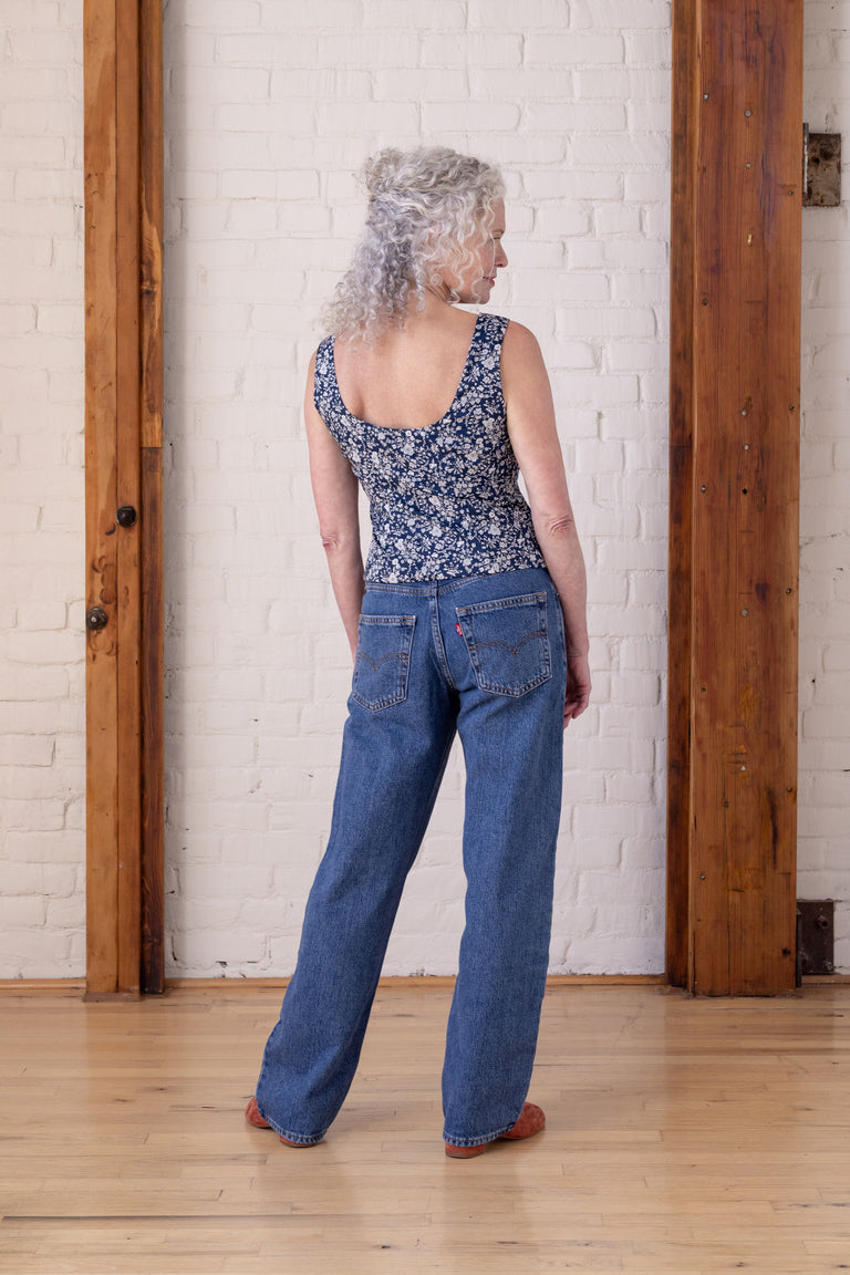 A woman with curly gray hair is shown from behind wearing a sleeveless blue and white floral print top and blue jeans, standing in a room with a white brick wall and wooden columns.
