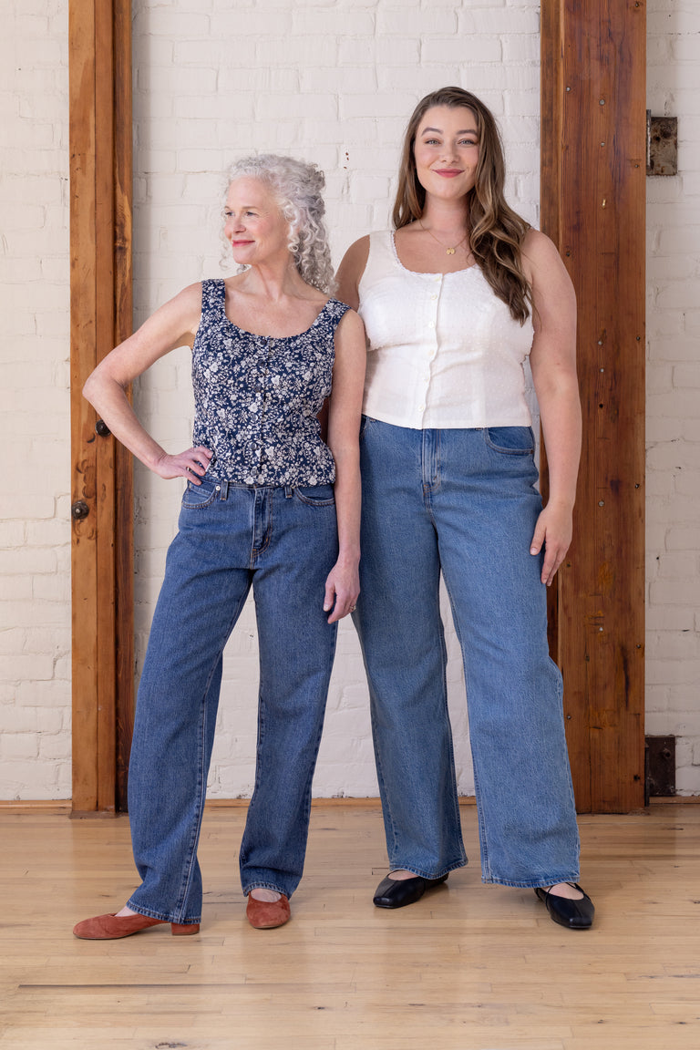 Two women modeling wide-leg blue jeans in a bright indoor studio with a white brick wall and wooden door frames. The woman on the left wears a navy floral tank top, and the woman on the right wears a white tank top.