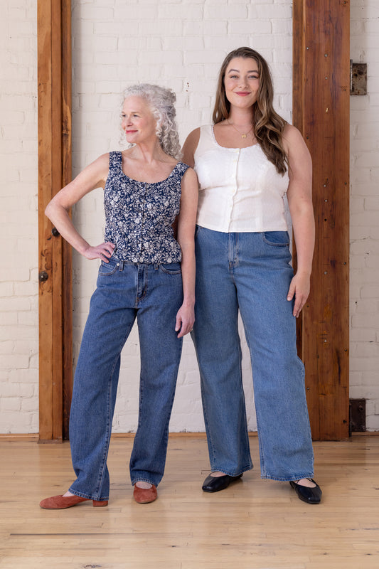 Two women modeling wide-leg blue jeans in a bright indoor studio with a white brick wall and wooden door frames. The woman on the left wears a navy floral tank top, and the woman on the right wears a white tank top.