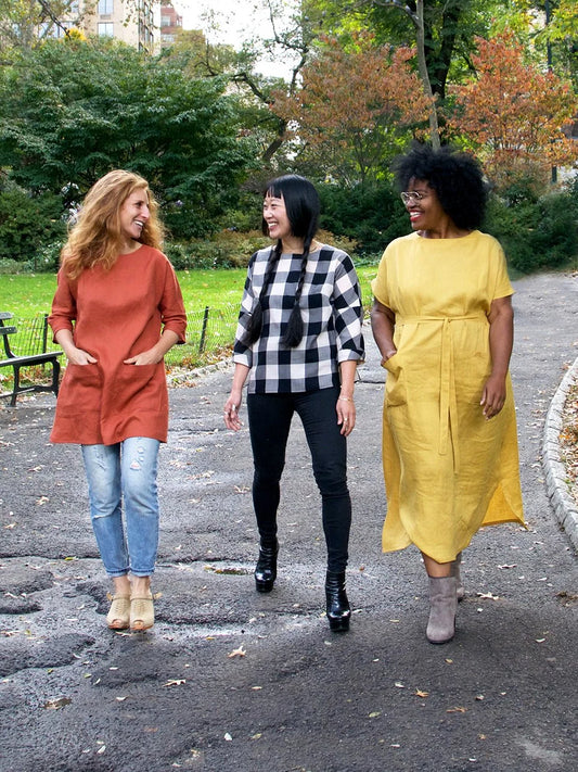 3 woman, one in a orange tunic, one in a checkers shirt and one in a yellow dress.  