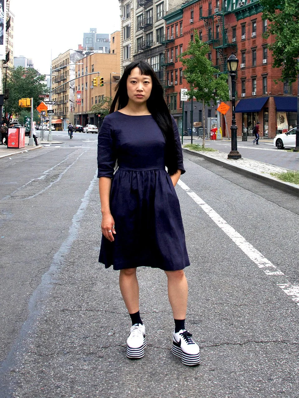 Woman in a navy dress standing on a city street with buildings and traffic lights in the background.