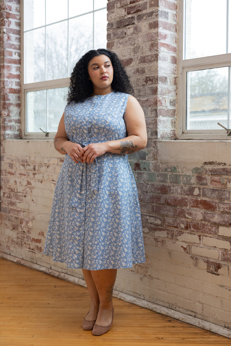 A person wearing a blue floral collarless shirt dress with a high neckline, sleeveless design, and waist tie.