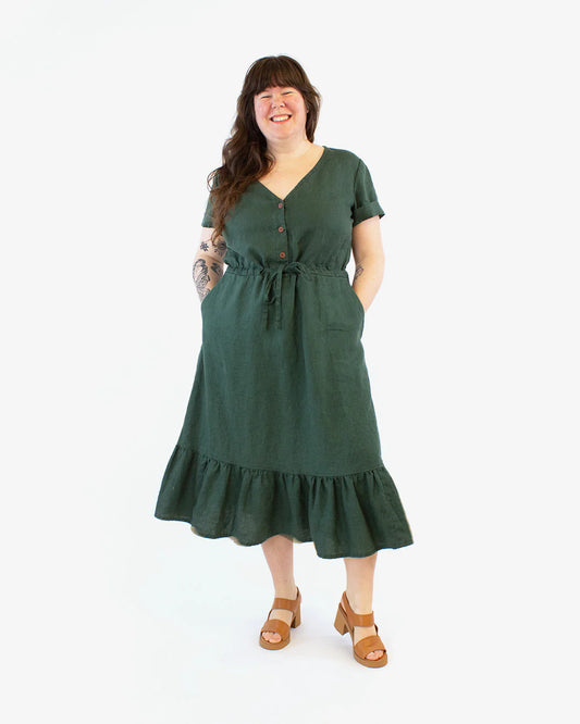 Full detail photo of woman wearing a green dress with a front button placket, drawstring waist and a bottom tier. Her hands are in pockets inserted into the skirt.  Photo taken against a white background.