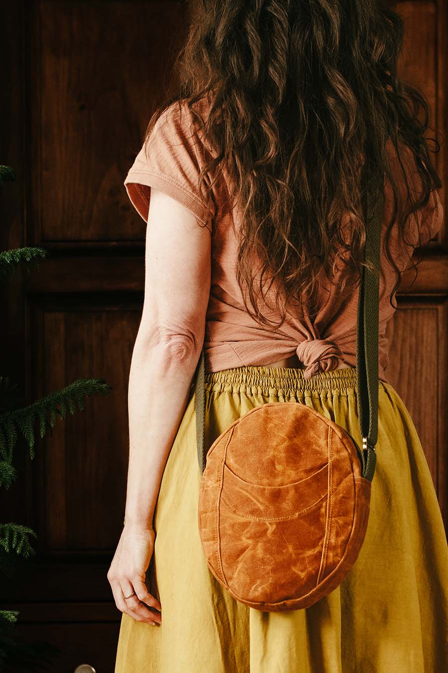 Person wearing a brown top and yellow skirt with a round brown leather bag over one shoulder, standing against a wooden paneled wall.