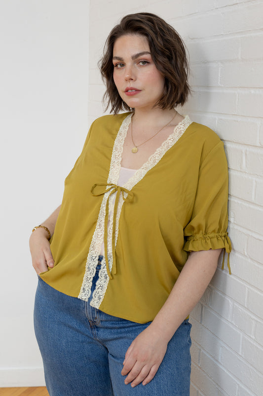 Woman wearing a mustard yellow blouse with lace details against a white brick wall.
