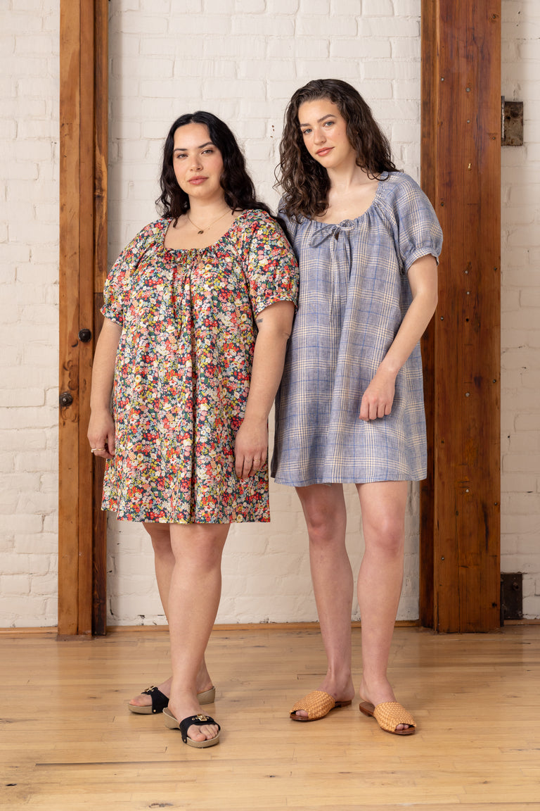 Two women standing side by side wearing short dresses against a white brick wall.