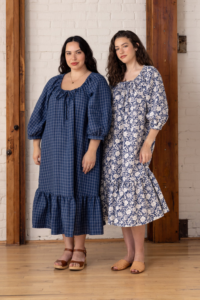 Two women wearing blue dresses standing in a room with wooden floors and white brick walls.