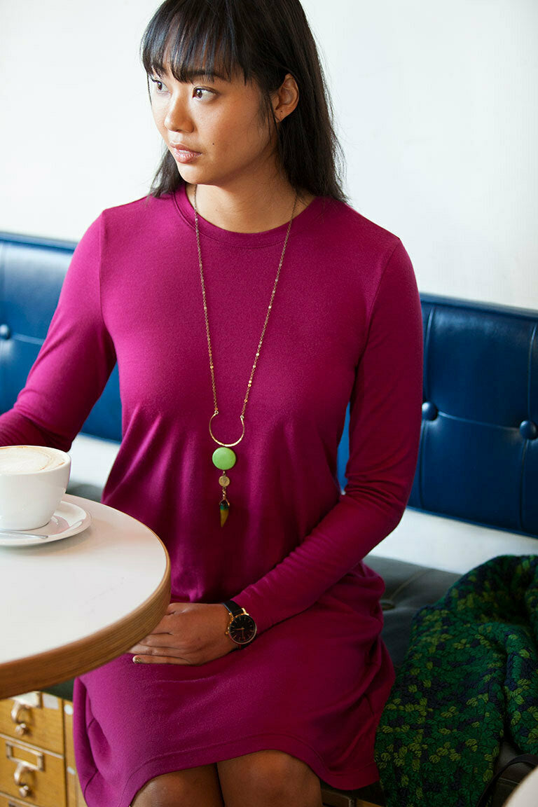 Woman in a magenta dress sitting at a table with a cup of coffee, wearing a long necklace.