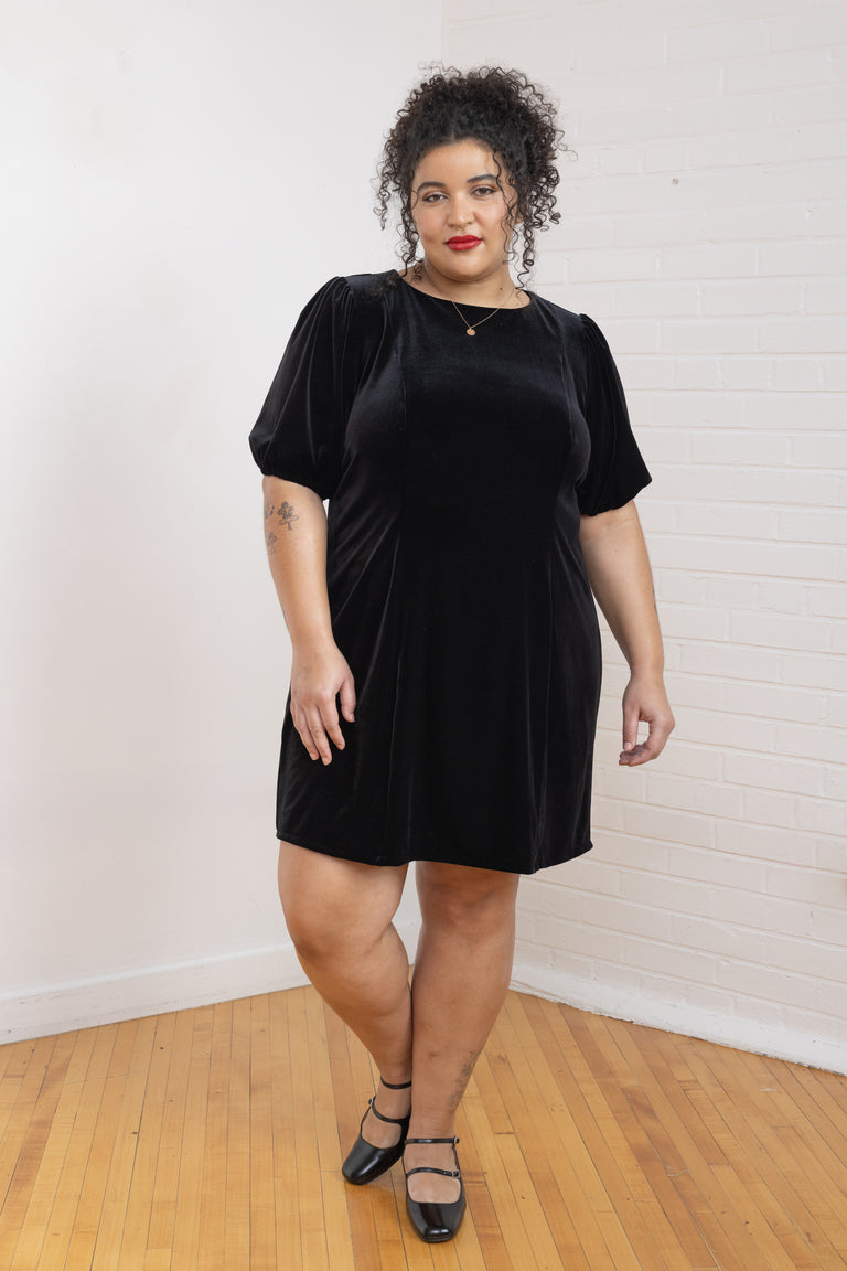 Woman wearing a black dress standing on a wooden floor with a white wall background