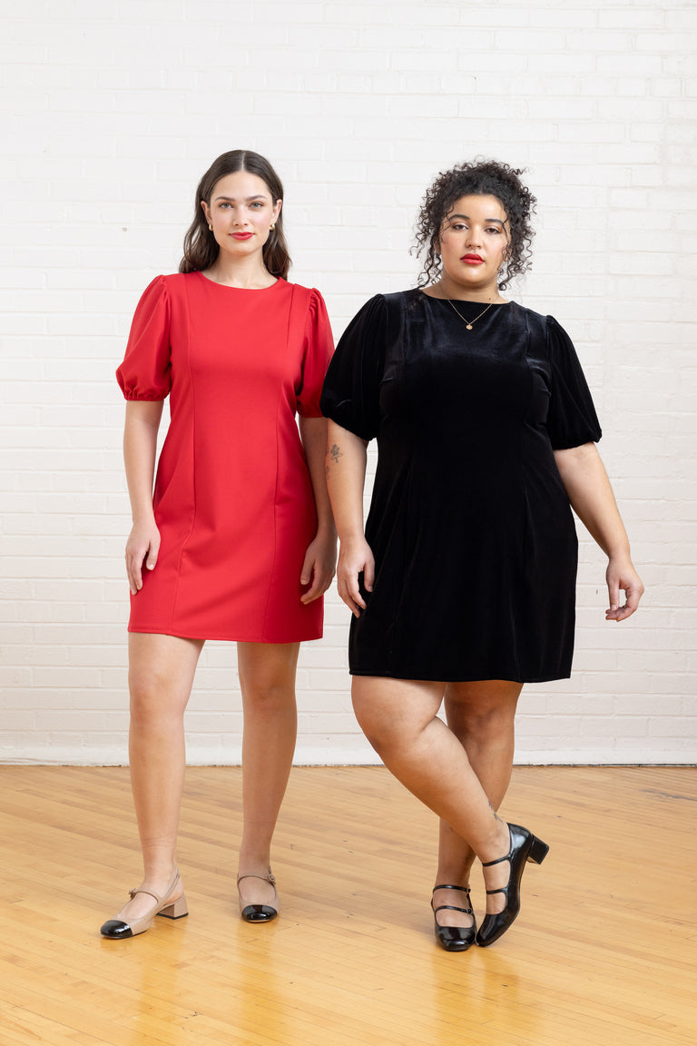 Two women wearing red and black dresses standing on a wooden floor with a white wall background.