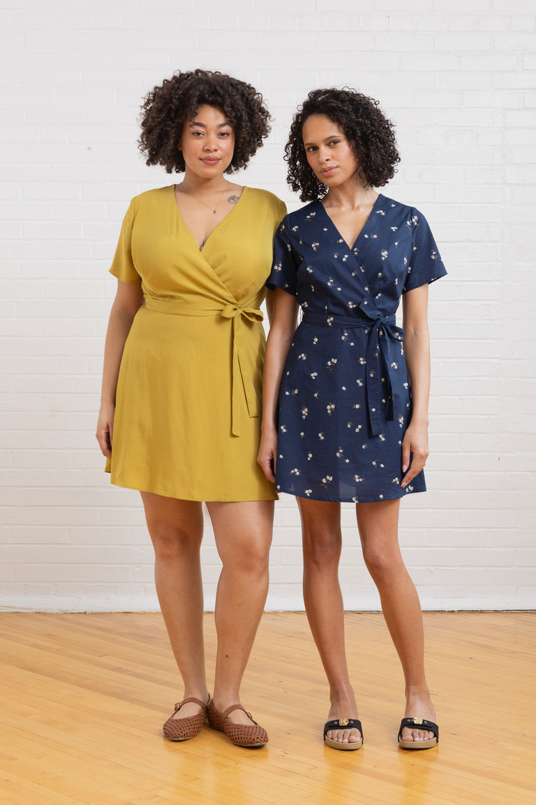 Two women wearing yellow and navy wrap dresses standing against a white brick wall.