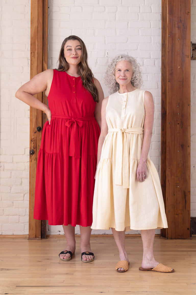 Two women modeling sleeveless midi dresses in a bright indoor studio with a white brick wall and wood trim. The woman on the left wears a red dress, and the woman on the right wears a light cream dress.