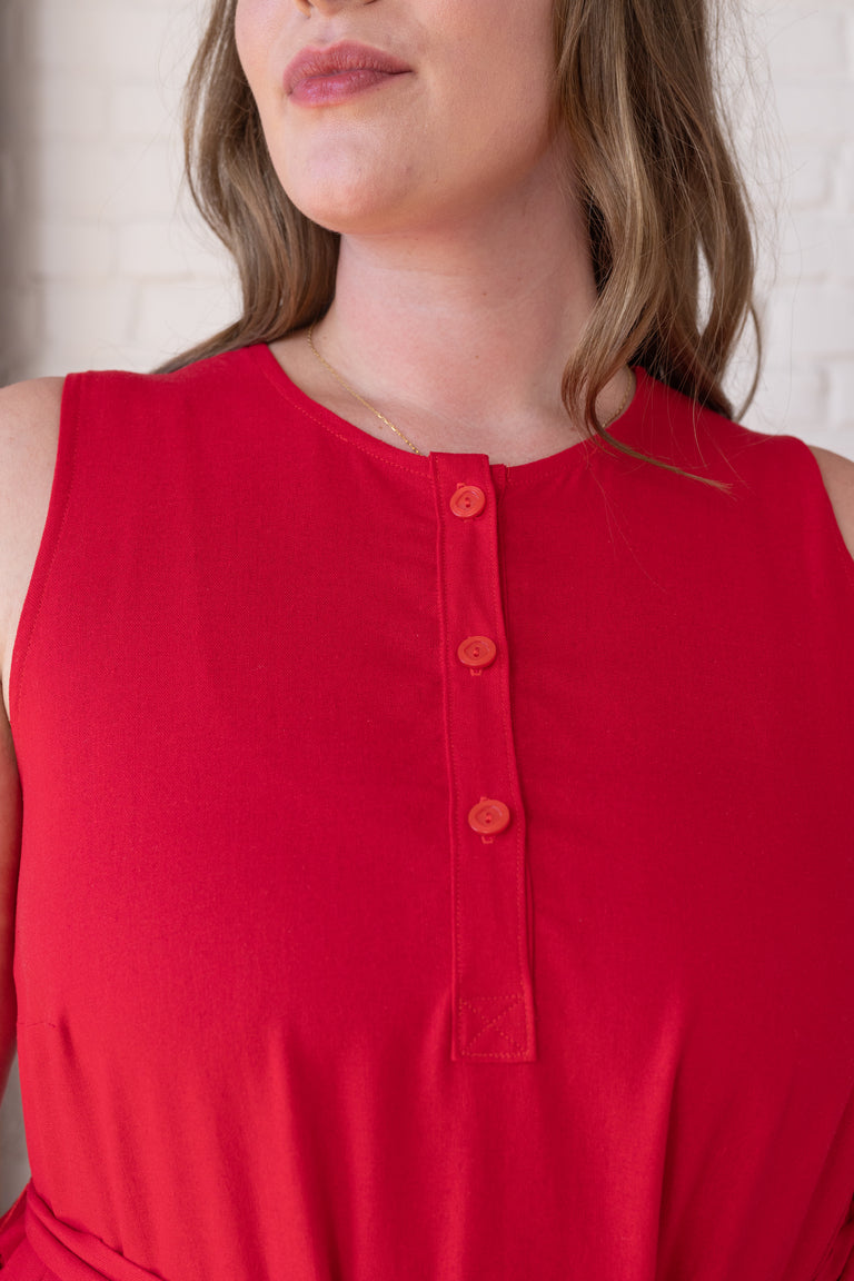 A close-up of a woman wearing a sleeveless red top with a three-button placket on the chest, set against a light brick wall background.