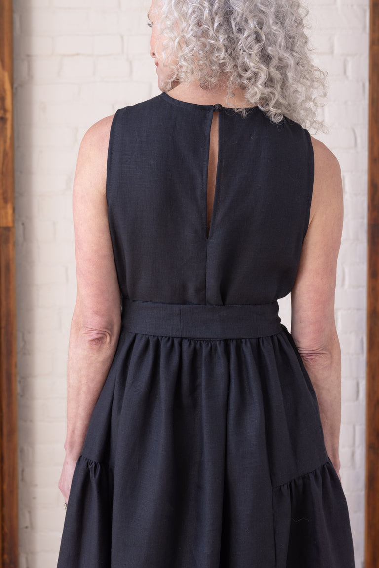 A woman shown from the back wearing a sleeveless black dress with a back slit and a defined waistband, standing against a white brick wall.