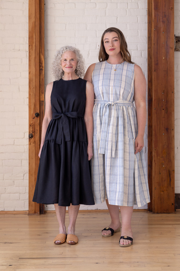 Two women modeling sleeveless midi dresses with tie waist details, standing in a bright room with a white brick wall and wooden columns. The woman on the left wears a solid black dress, and the woman on the right wears a light blue and white plaid dress.