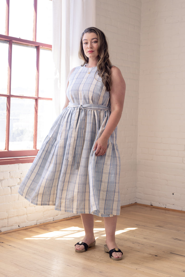 A woman wearing a sleeveless, knee-length plaid dress in light blue, beige, and white, standing in a bright room with white brick walls and a wooden floor.