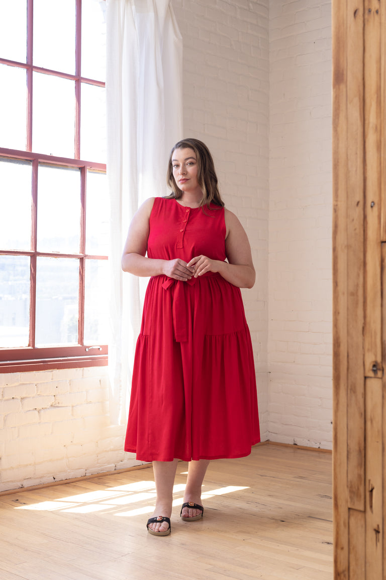 A woman wearing a sleeveless red tiered midi dress with a buttoned neckline, standing in a bright room with white brick walls, a large window, and light wood floors.