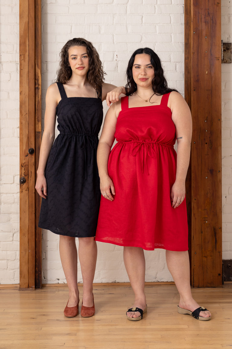 Two women standing side by side, one in a black dress and the other in a red dress, against a white brick wall.