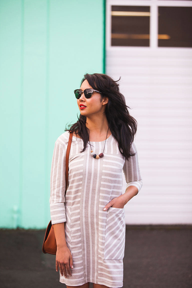 Woman wearing a white dress with stripes, sunglasses, and a brown bag against a teal and white wall.
