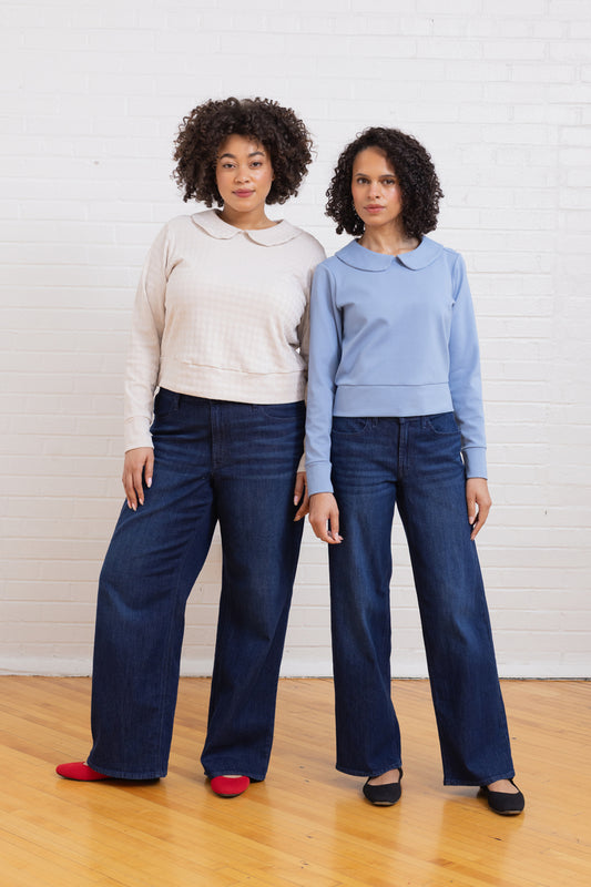 Two women standing side by side wearing matching outfits of light-colored tops and dark blue jeans against a white brick wall.
